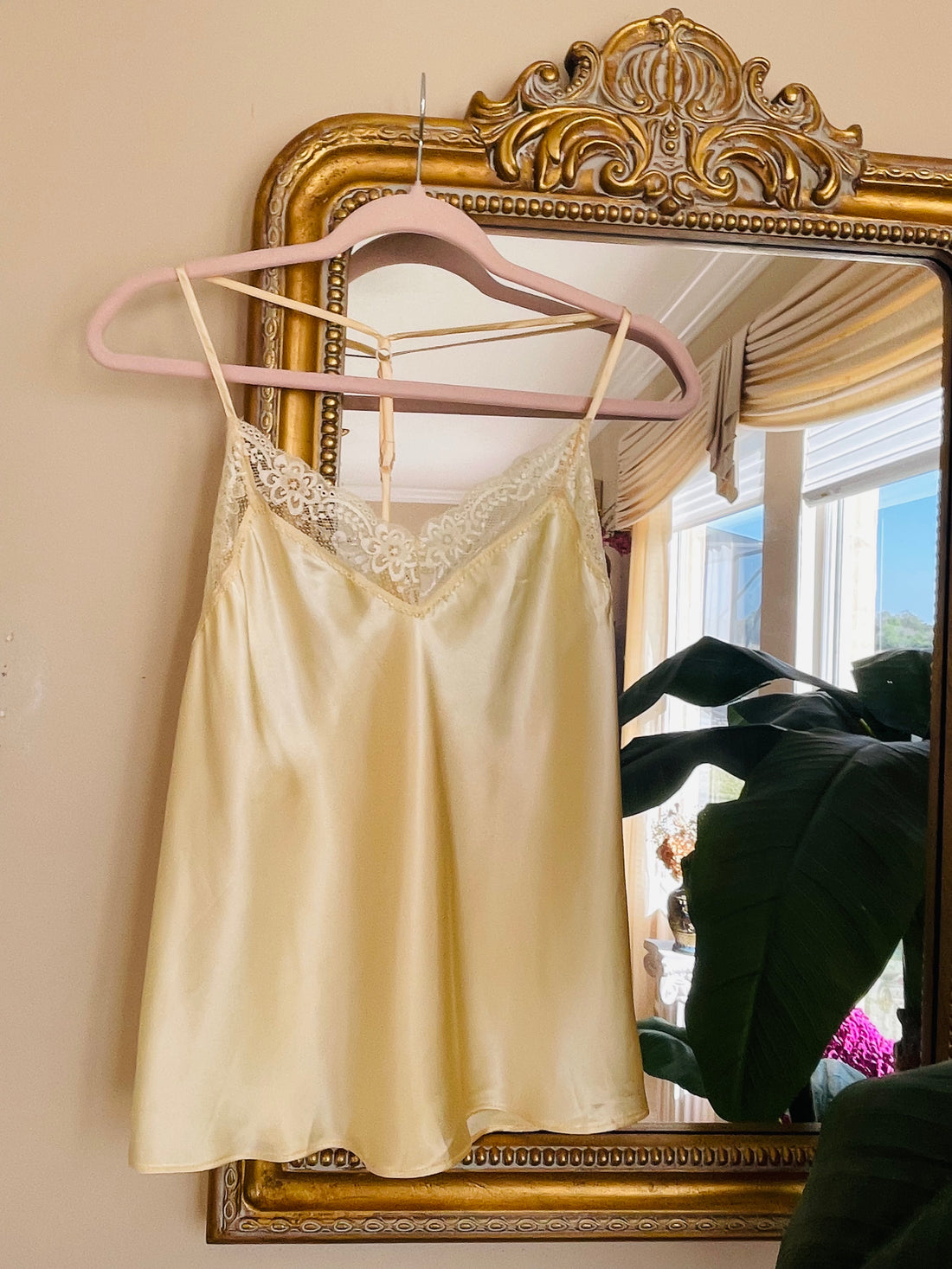 Beige satin camisole with lace trim on a pink hanger in front of a decorative mirror.